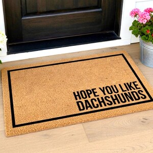 May include: A rectangular coir doormat with a black border and the text "HOPE YOU LIKE DACHSHUNDS" in black. The mat is placed in front of a dark brown door. The mat is made of natural coir fiber.
