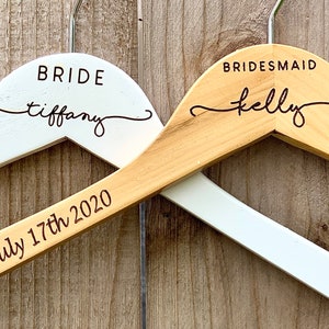 May include: Two wooden hangers with engraved text. The top hanger is white with the text "BRIDE tiffany". The bottom hanger is natural wood with the text "BRIDESMAID kelly" and "July 17th 2020".