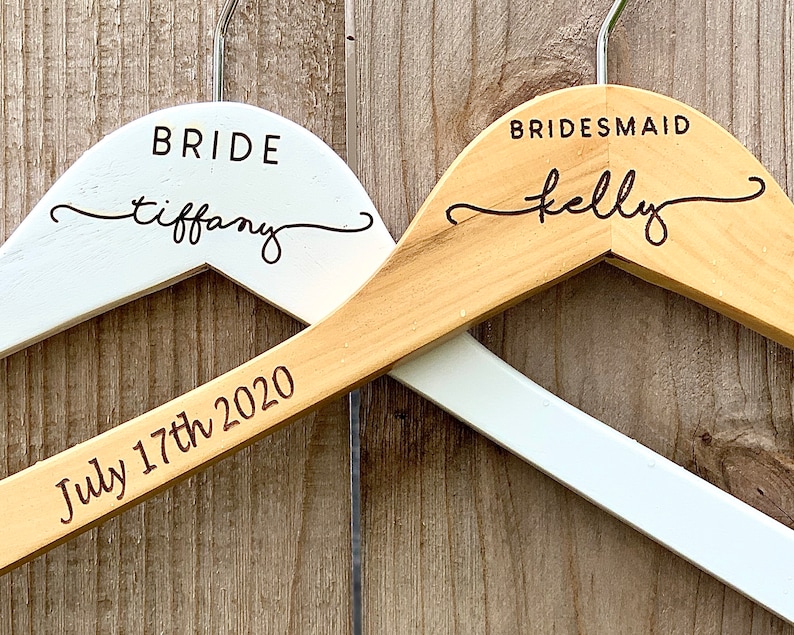 May include: Two wooden hangers with engraved text. The top hanger is white with the text "BRIDE tiffany". The bottom hanger is natural wood with the text "BRIDESMAID kelly" and "July 17th 2020".