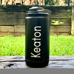 May include: Black insulated water bottle with a flip-top lid. The word "Keton" is printed vertically in white on the front of the bottle. The bottle is sitting on a wooden surface with green grass and a wooden fence in the background.