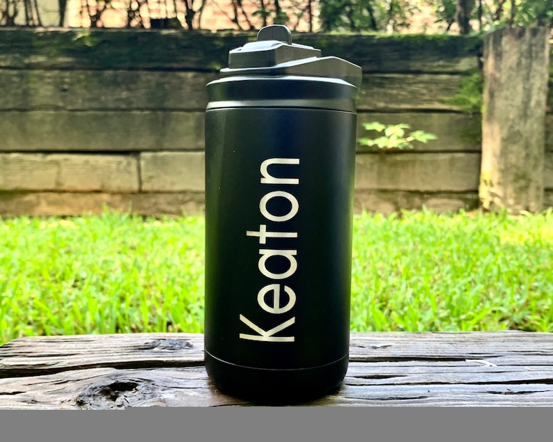 May include: Black insulated water bottle with a flip-top lid. The word "Keton" is printed vertically in white on the front of the bottle. The bottle is sitting on a wooden surface with green grass and a wooden fence in the background.