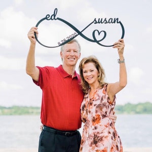May include: A couple hold a black metal infinity symbol with the words "ed" and "susan" on either side, and the date "8.28.2020" inscribed in the centre. A heart is in the centre of the infinity symbol.
