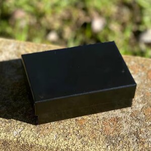 May include: A small, rectangular black box with a glossy finish. The box is sitting on a rough, textured, light brown surface, with a blurred green background.