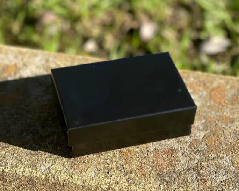 May include: A small, rectangular black box with a glossy finish. The box is sitting on a rough, textured, light brown surface, with a blurred green background.