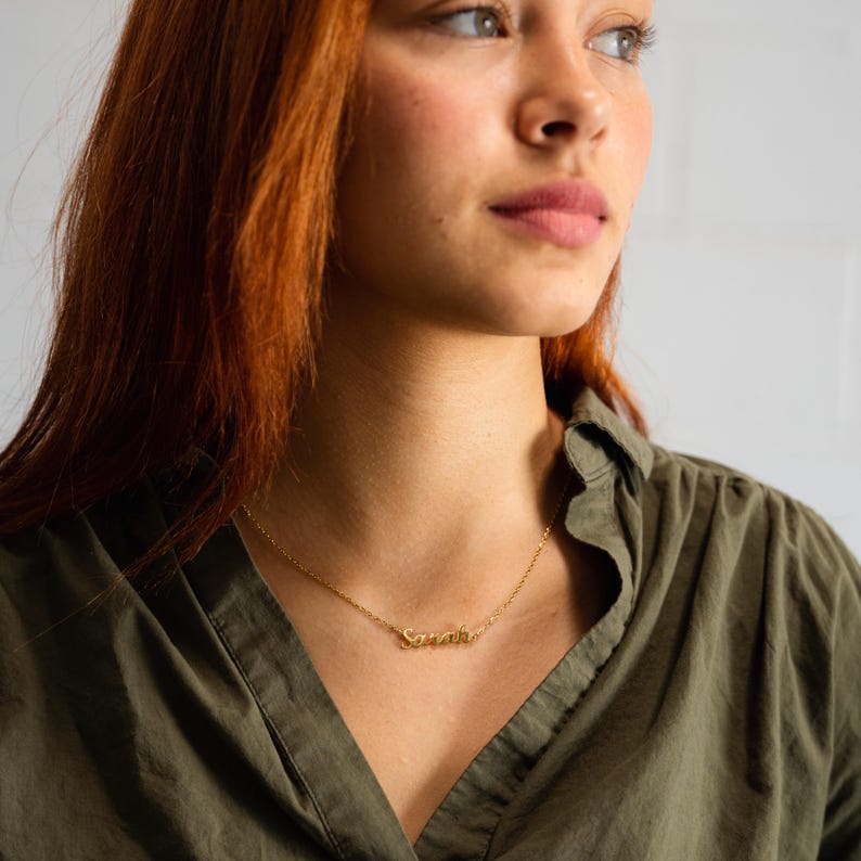 May include: A gold-colored necklace with the name "Sarah" in cursive script. The necklace is worn against a green shirt. The chain is delicate and the pendant is small. The background is a plain white wall.