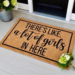 May include: Rectangular doormat with the text "THERE'S LIKE, a lot of girls IN HERE" in black script. The mat is brown with a black border and is placed on a light gray surface. A bouquet of white and green flowers is on the right.