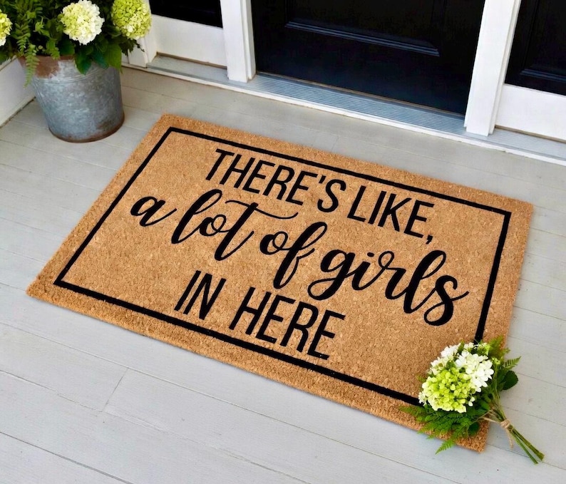 May include: Rectangular doormat with the text "THERE'S LIKE, a lot of girls IN HERE" in black script. The mat is brown with a black border and is placed on a light gray surface. A bouquet of white and green flowers is on the right.