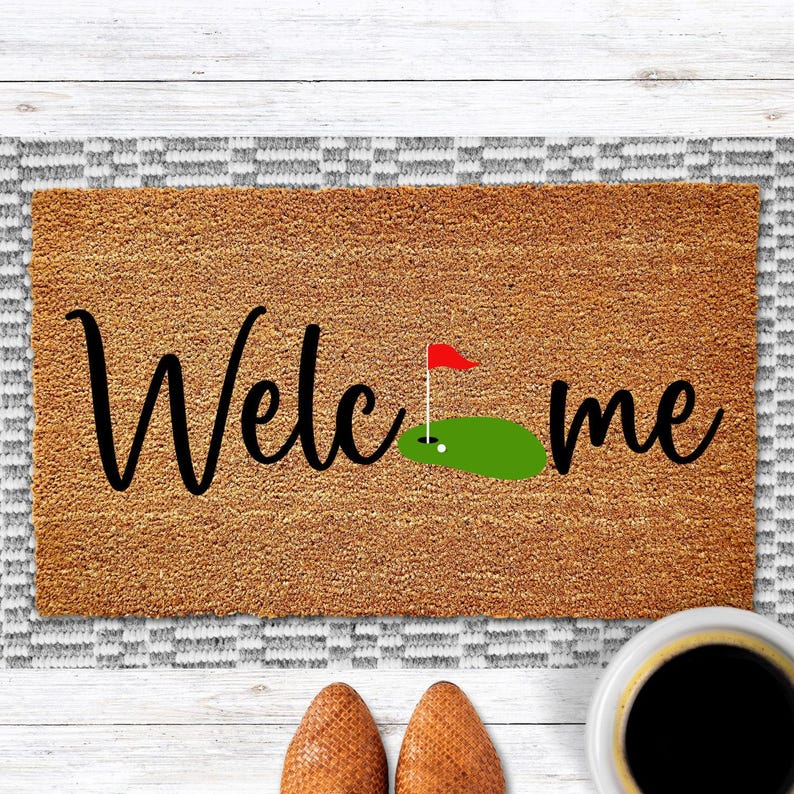 May include: A brown rectangular doormat with the word "Welcome" in black script, a golf green graphic, and a red flag. The mat has a textured surface and is placed on a patterned rug. A pair of brown shoes and a cup of coffee are in the foreground.