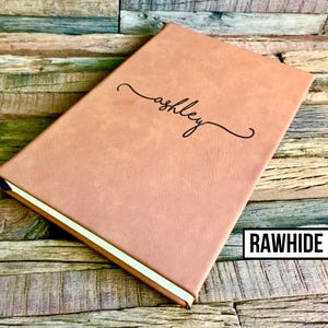 May include: A personalized, light brown journal with the name "ashley" in cursive script. The journal has a gold-colored edge and the text "RAWHIDE 2" in a white box.