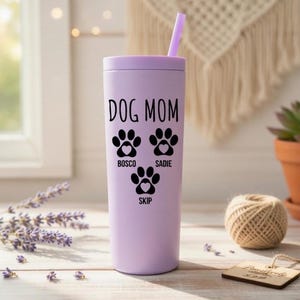 K&ouml;nnte beinhalten: Ein helllila Becher mit passendem Deckel und Strohhalm. Der Becher tr&auml;gt die schwarzen Worte "DOG MOM" sowie drei Pfotenabdr&uuml;cke und die Namen "Bosco", "Sadie" und "Skip". Er steht auf einer Holzoberfl&auml;che.