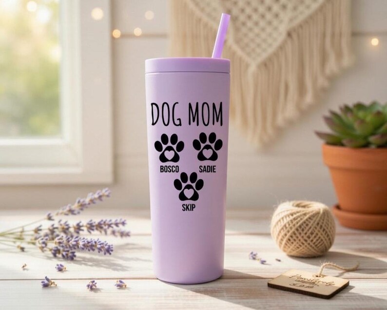 K&ouml;nnte beinhalten: Ein helllila Becher mit passendem Deckel und Strohhalm. Der Becher tr&auml;gt die schwarzen Worte "DOG MOM" sowie drei Pfotenabdr&uuml;cke und die Namen "Bosco", "Sadie" und "Skip". Er steht auf einer Holzoberfl&auml;che.