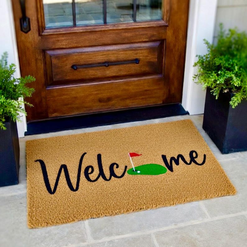 May include: A rectangular doormat with the word "Welcome" in black script. A green golf course graphic with a red flag is next to the word "Welcome". The mat is tan and sits in front of a wooden door.