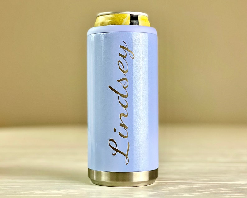 May include: A pale blue can cooler with the name "Lindsey" in gold script. The cooler has silver rims and is designed to hold a standard can. A yellow and white can is inside the cooler.