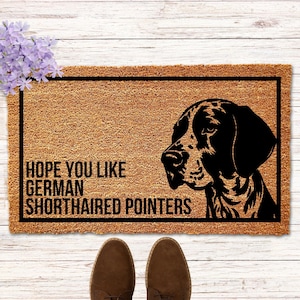 May include: A rectangular doormat with a black border and the text "HOPE YOU LIKE GERMAN SHORTHAIRED POINTERS". The mat features a black silhouette of a German Shorthaired Pointer dog. Brown shoes are at the bottom.