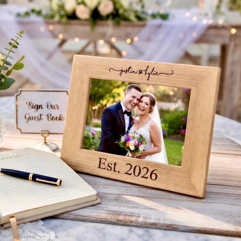 May include: A light-colored wooden picture frame with a wedding photo of a smiling couple. The frame is engraved with the names "justin & kylie" and the date "Est. 2026". A sign that says "Sign Our Guest Book" is visible.