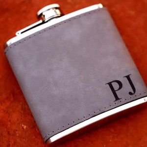 May include: A gray leather-covered flask with a silver lid and a silver rim. The flask has the initials "PJ" engraved on the front.