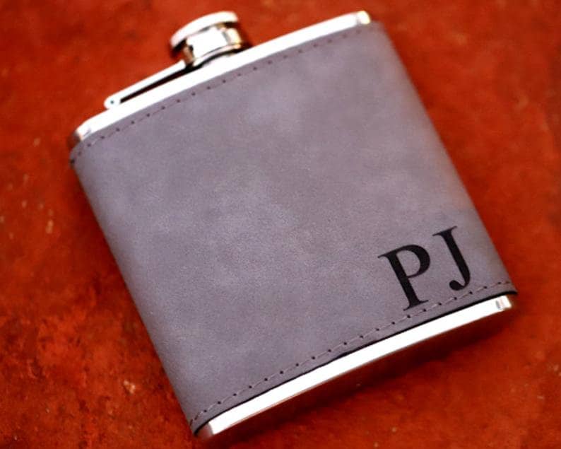 May include: A gray leather-covered flask with a silver lid and a silver rim. The flask has the initials "PJ" engraved on the front.
