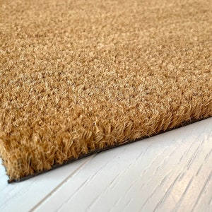 May include: Close-up of a rectangular doormat made of natural brown coir fibers. The mat has a dark border and is placed on a white wooden surface. The coir fibers are densely packed, creating a textured surface.