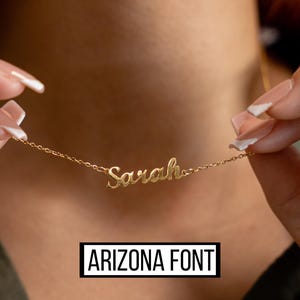 May include: A gold-colored necklace with the name "Sarah" in cursive script. The necklace is held up by a person. The text "ARIZONA FONT" is displayed in a black rectangle at the bottom of the image.
