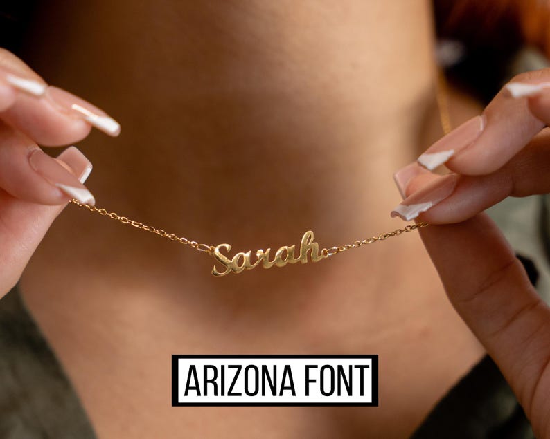 May include: A gold-colored necklace with the name "Sarah" in cursive script. The necklace is held up by a person. The text "ARIZONA FONT" is displayed in a black rectangle at the bottom of the image.
