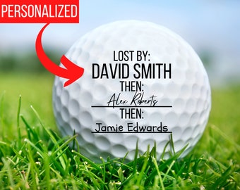 May include: A white golf ball with black text that reads "LOST BY: DAVID SMITH THEN: Alex Roberts THEN: Jamie Edwards". The golf ball is on a green grassy surface.