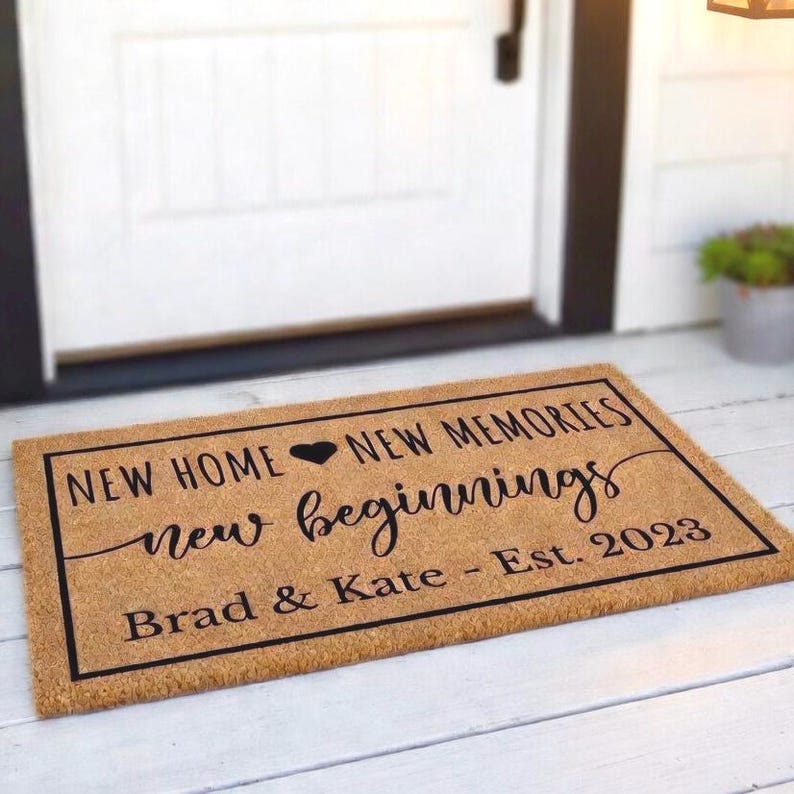 May include: A rectangular doormat with a black border and text. The text reads "NEW HOME, NEW MEMORIES, new beginnings" with a heart symbol, and "Brad & Kate - Est. 2023". The mat is a natural brown color and sits in front of a white door.