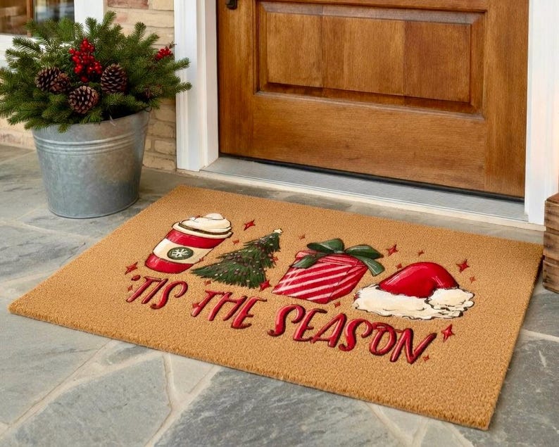 May include: A rectangular doormat with a festive Christmas design. It features a red and white coffee cup, a green Christmas tree, a wrapped gift, and a Santa hat. The words "'Tis the Season" are written in red.