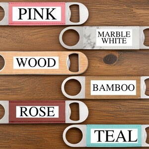 May include: A collection of bottle openers with stainless steel bodies and color-coded inserts. The inserts display the words "PINK", "MARBLE WHITE", "WOOD", "BAMBOO", "ROSE", and "TEAL". The openers are arranged on a wooden surface.