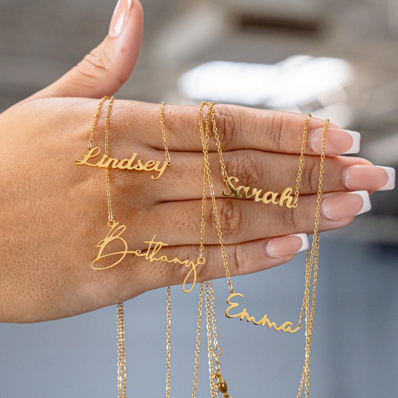May include: Gold-colored name necklaces spelling out "Lindsey", "Bethany", "Sarah", and "Emma" in a cursive font. The necklaces are displayed against a neutral background.