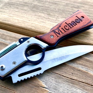 Puede incluir: Un cuchillo de bolsillo plateado y negro con un mango de madera grabado con el nombre "Michael". El cuchillo tiene un encendedor incorporado y una hoja dentada.