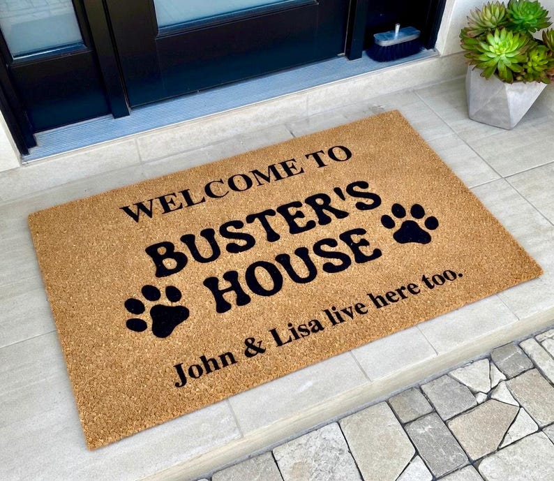 May include: A rectangular doormat made of natural coir fiber. The mat features the text "WELCOME TO BUSTER'S HOUSE" in black, along with paw prints. The names "John & Lisa" are also printed on the mat.