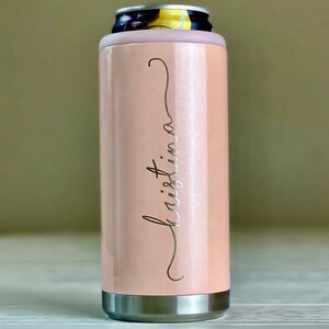May include: A pale pink insulated can cooler with a silver base and rim. The name "Kristina" is written in a cursive font on the side. The top of the cooler has a light purple rim to hold a can.
