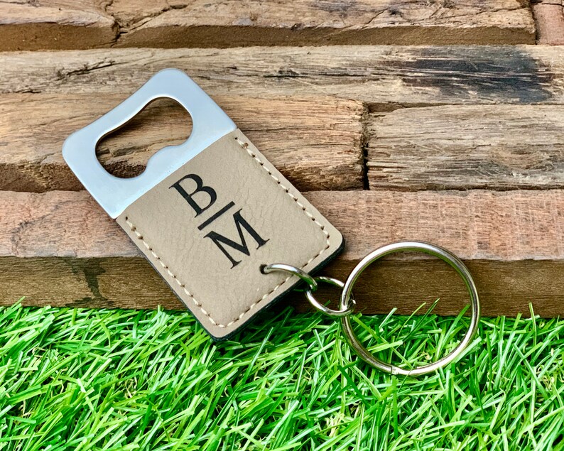 May include: A brown leather bottle opener with a silver metal opener and a keychain ring. The bottle opener is engraved with the initials "B|M".