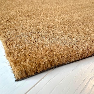 May include: A rectangular doormat made of natural tan coir fibres with a dense, coarse, and bristly texture. The mat features a thick, fibrous surface layer attached to a dark, solid base, showing a sharp corner and straight edges.