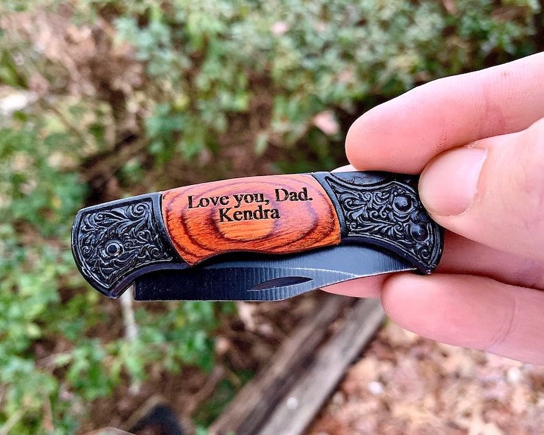 May include: A small pocket knife with a wooden handle engraved with the words "Love you, Dad. Kendra". The handle is brown with black accents. The blade is black and partially open.