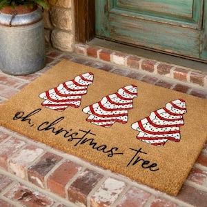 May include: A rectangular doormat made of natural fibers. It features three Christmas tree designs in red and white, with a striped pattern. The words "oh, christmas tree" are written in a cursive font.