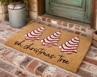 May include: A rectangular doormat made of natural fibres. It features three Christmas tree designs in red and white, with a striped pattern. The words "oh, christmas tree" are written in a cursive font.