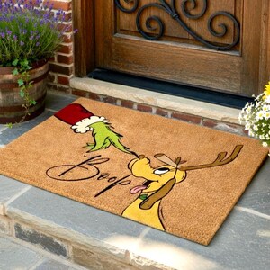 May include: A rectangular doormat featuring a cartoon dog with antlers and a green hand reaching out from a red hat. The word "Boop" is written in a script font. The doormat is made of a natural fibre material.