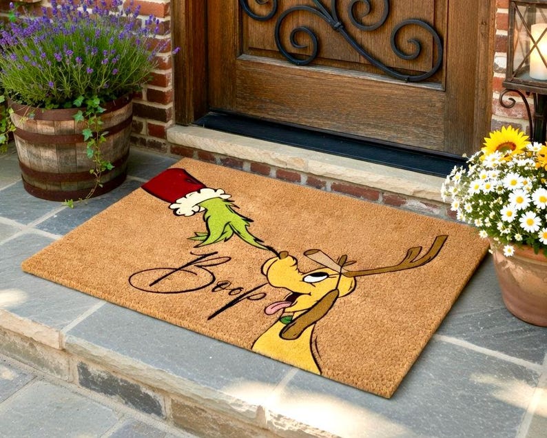 May include: A rectangular doormat featuring a cartoon dog with antlers and a green hand reaching out from a red hat. The word "Boop" is written in a script font. The doormat is made of a natural fibre material.