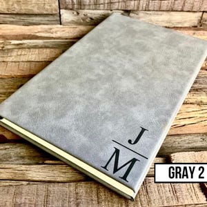 May include: A gray journal with a textured cover and gold-colored edges. The initials "JM" are printed in black on the front cover. The text "GRAY 2" is in a white box in the lower right corner. The journal is on a wooden surface.