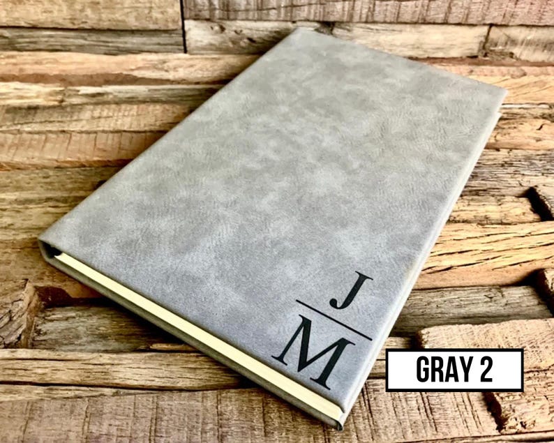 May include: A gray journal with a textured cover and gold-colored edges. The initials "JM" are printed in black on the front cover. The text "GRAY 2" is in a white box in the lower right corner. The journal is on a wooden surface.