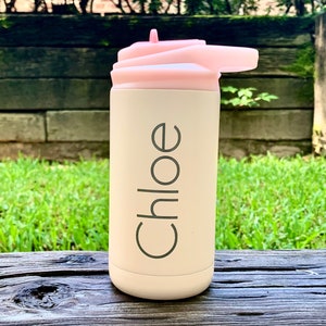 May include: A light beige water bottle with a pink lid and a flip-top spout. The name "Chloe" is printed vertically in a dark gray font on the bottle. The bottle is sitting on a wooden surface with a green grassy background.