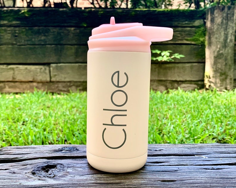 May include: A light beige water bottle with a pink lid and a flip-top spout. The name "Chloe" is printed vertically in a dark gray font on the bottle. The bottle is sitting on a wooden surface with a green grassy background.