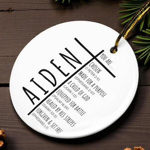 May include: A white ceramic ornament with the name "AIDEN" in large black letters. The ornament features a cross design with inspirational text, including "CHOSEN" and "MADE FOR A PURPOSE". A gold string is attached for hanging.