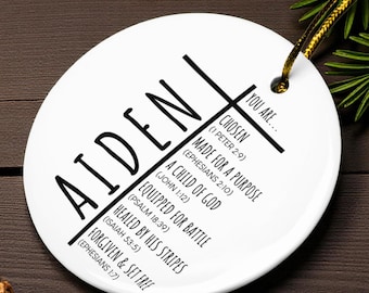 May include: A white ceramic ornament with the name "AIDEN" in large black letters. The ornament features a cross design with inspirational text, including "CHOSEN" and "MADE FOR A PURPOSE". A gold string is attached for hanging.