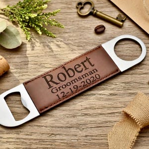 May include: A brown leather and stainless steel bottle opener with the name "Robert", the word "Groomsman", and the date "12-19-2020" engraved on the leather. The opener is on a wooden surface with decorative elements.