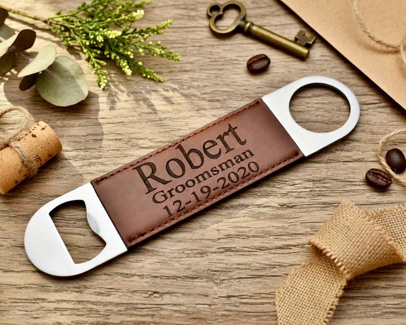 May include: A brown leather and stainless steel bottle opener with the name "Robert", the word "Groomsman", and the date "12-19-2020" engraved on the leather. The opener is on a wooden surface with decorative elements.