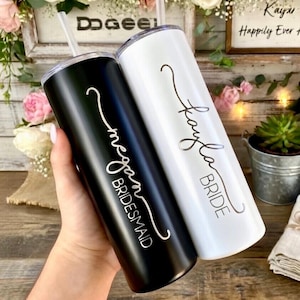 May include: Two stainless steel tumblers, one black and one white, held by a hand. The black tumbler says "Megan Bridesmaid" in white script. The white tumbler says "Kayla Bride" in silver script. Both have clear straws.