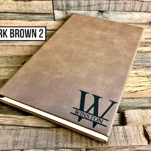 May include: A dark brown journal with the text "WINSTON" and a stylized "W" on the cover. The journal has a gold-colored edge and the text "DARK BROWN 2" in a black rectangle.