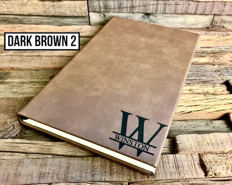 May include: A dark brown journal with the text "WINSTON" and a stylized "W" on the cover. The journal has a gold-colored edge and the text "DARK BROWN 2" in a black rectangle.